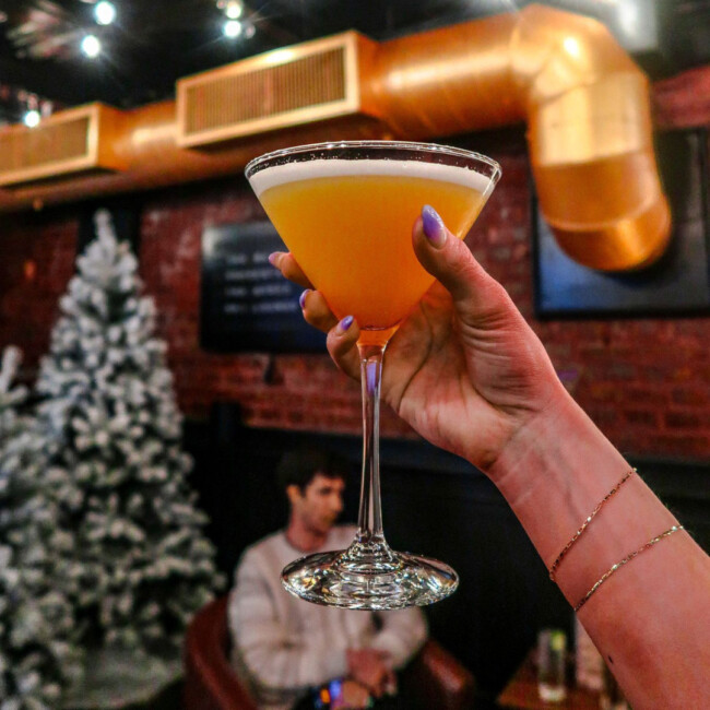 Hand with purple nails holding an orange cocktail in front of a Christmas tree